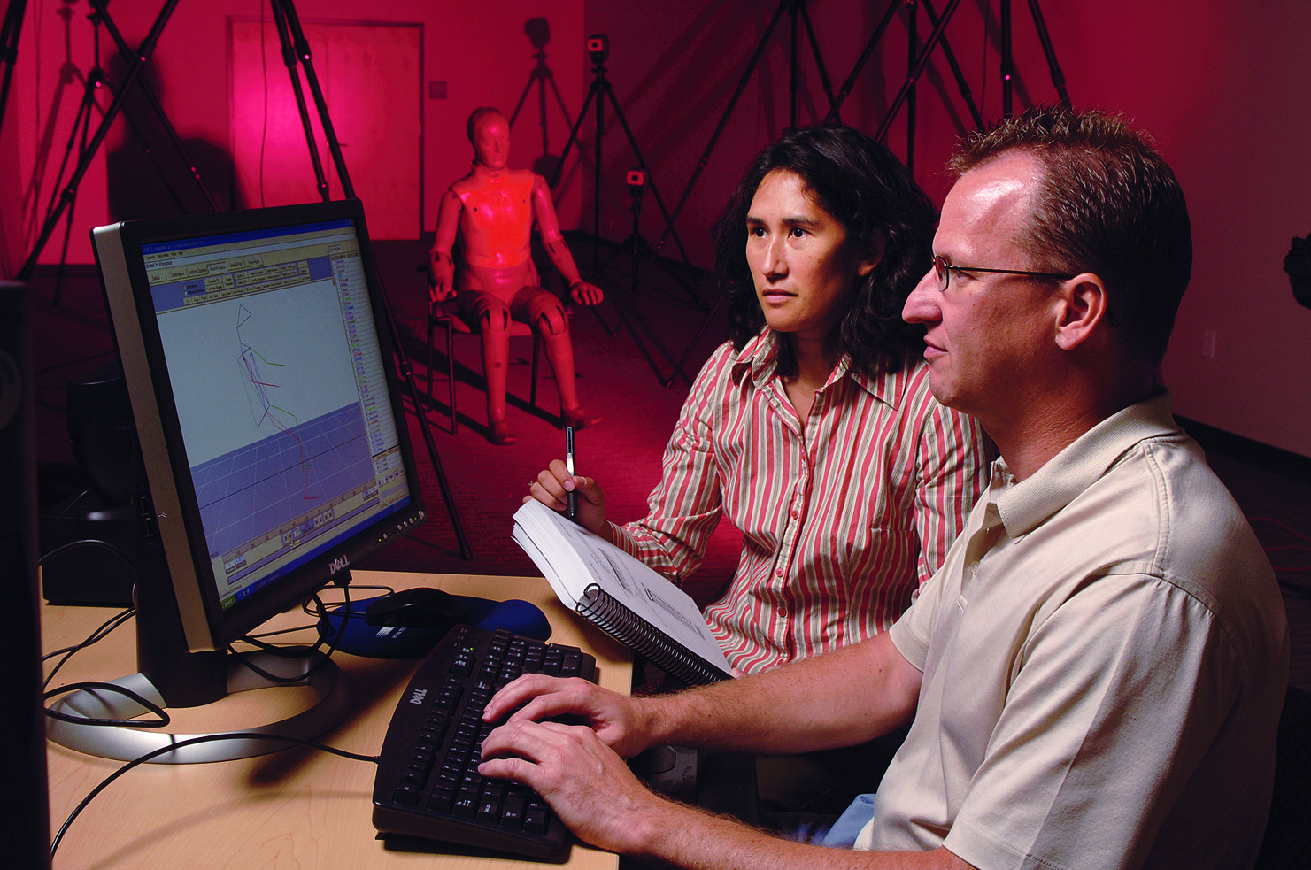 A man sits at a computer while a woman takes notes of what is on the screen