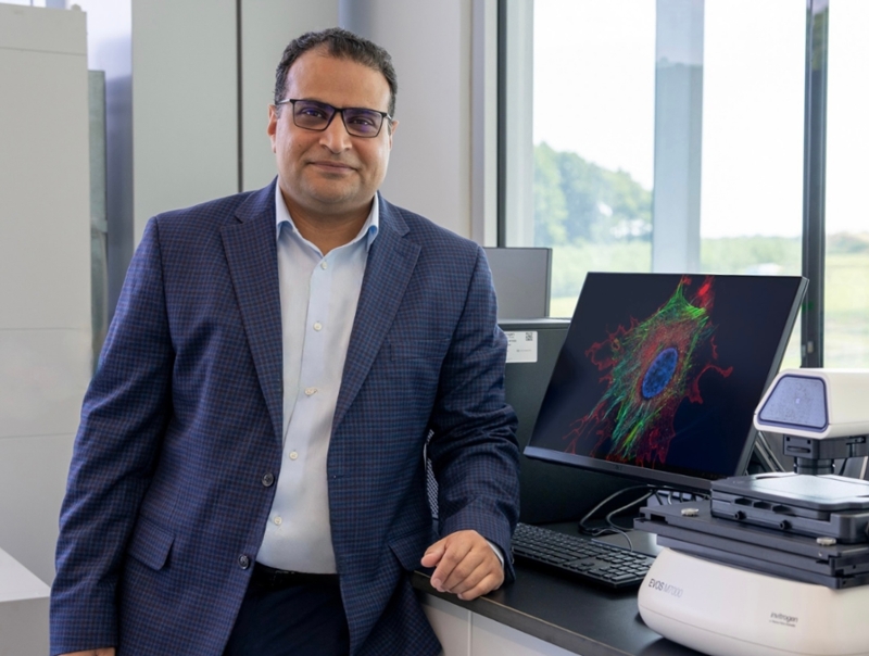 Mohamed Abouelkhair standing in a laboratory beside scientific imaging equipment and a computer displaying a microscope image.