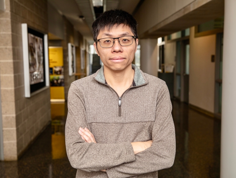 Chen Shen wearing glasses and a sweater, standing with arms crossed in a university hallway.