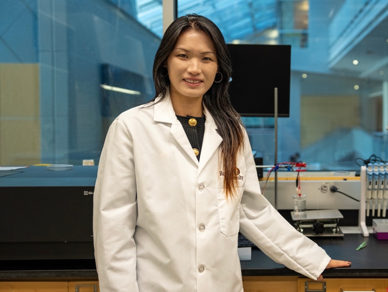 Dongmei Dong wearing a white lab coat stands beside a laboratory workbench with scientific instruments, pipettes and computer monitors in a research lab.