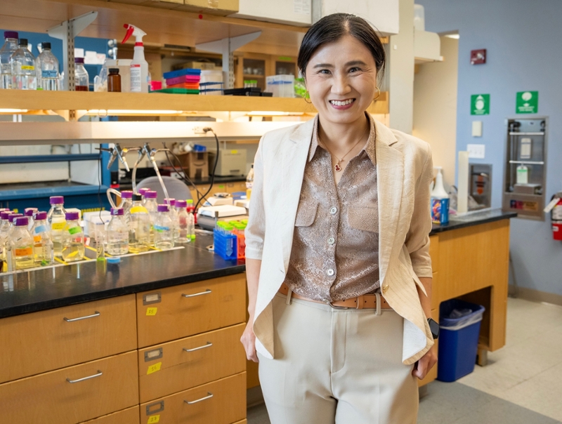 JiaBei Lin wearing a tan suit stands in a research lab with a workbench in the background.
