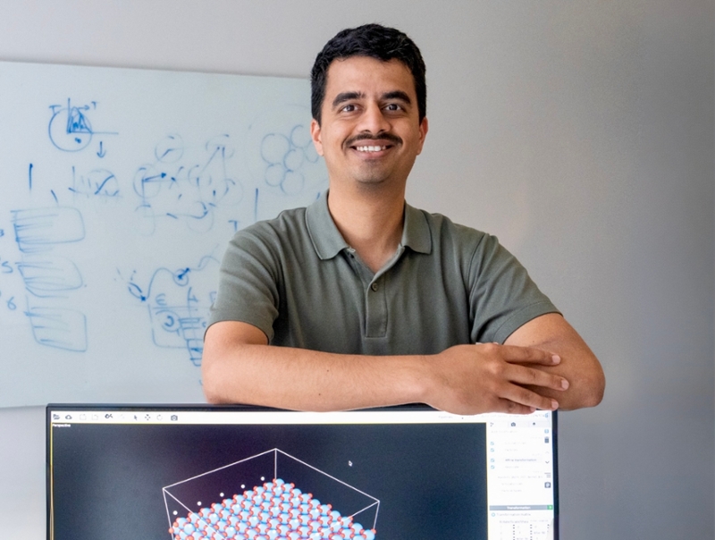 Aditya Lele, wearing a green polo shirt, stands in the foreground of a modern academic building hallway.