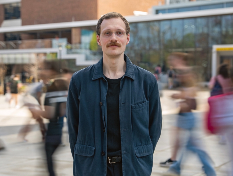 Maxi Heitmayer stands outdoors on a university campus walkway, wearing a dark jacket and shirt, with people walking past in motion blur behind them.