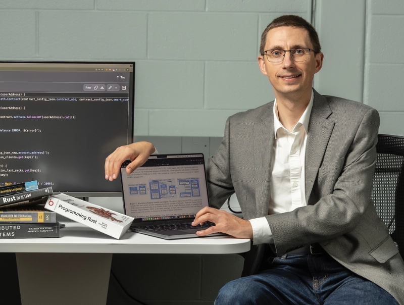 Nick Ivanov sits next to computer monitors with coding displayed.