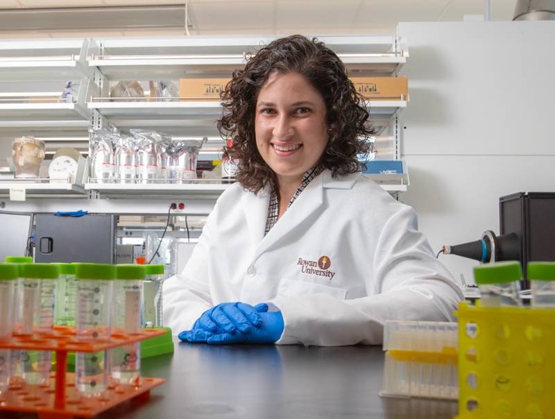 Researcher Rachel Riley in a lab wearing a white coat and gloves, seated beside racks of test tubes and lab equipment at Rowan University.