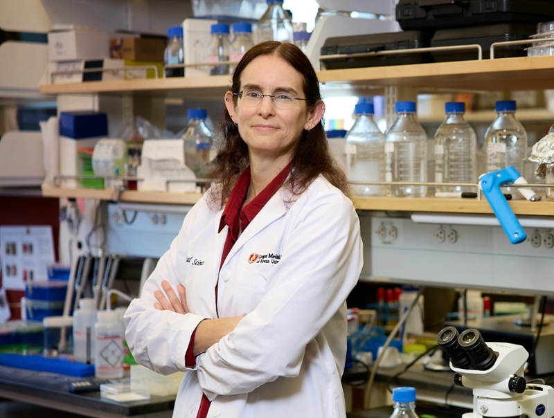 Susy Kohout of Cooper Medical School at Rowan University stands in a laboratory wearing a white lab coat with arms crossed. A microscope sits on the workbench in front, surrounded by bottles, pipettes, and racks of lab supplies. 